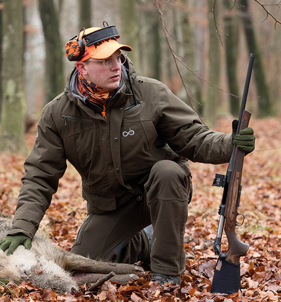 Ein Jäger im herbstlichen Laubwald mit orangener Käppi. Seine rechte Hand liegt auf einem erlegten Wilschwein, in der rechten Hand hält er sein Gewehr.