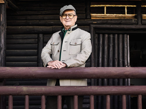 Ein alter Mann steht posierend in jagdsicher Kleidung vor einer Blockhütte und schaut forsch in die Kamera