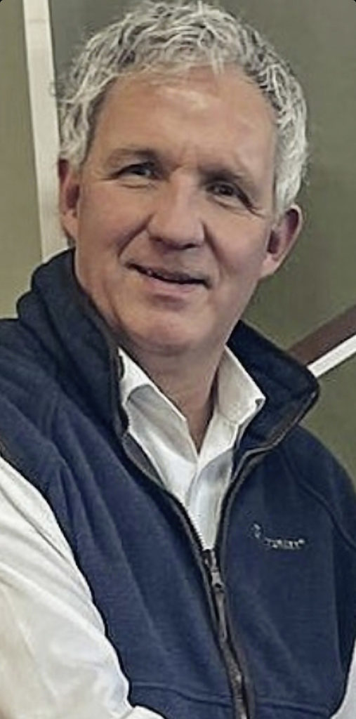 Ein Mann in hellem Hemd mit dunkelblauer Weste und grauem Haar lächelt in die Kamera